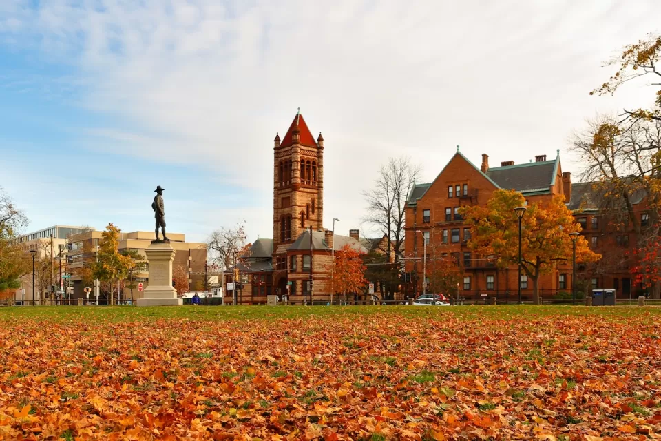 John Bridge Monument (also known as The Puritan), in the northeast corner of the Cambridge Common in Cambridge, Massachusetts with fall foliage on ground.
